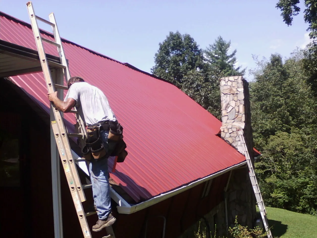 Roofer on a ladder beside a red metal roof and stone chimney — Soffit & Fascia Repair by Roofing Brothers in Slaughters